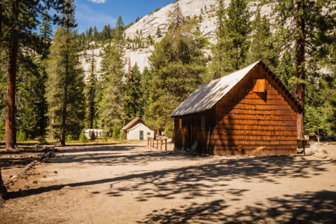 A shingle-sided camp building in the clearing, beneath the white granite wall above the Merced River canyon.