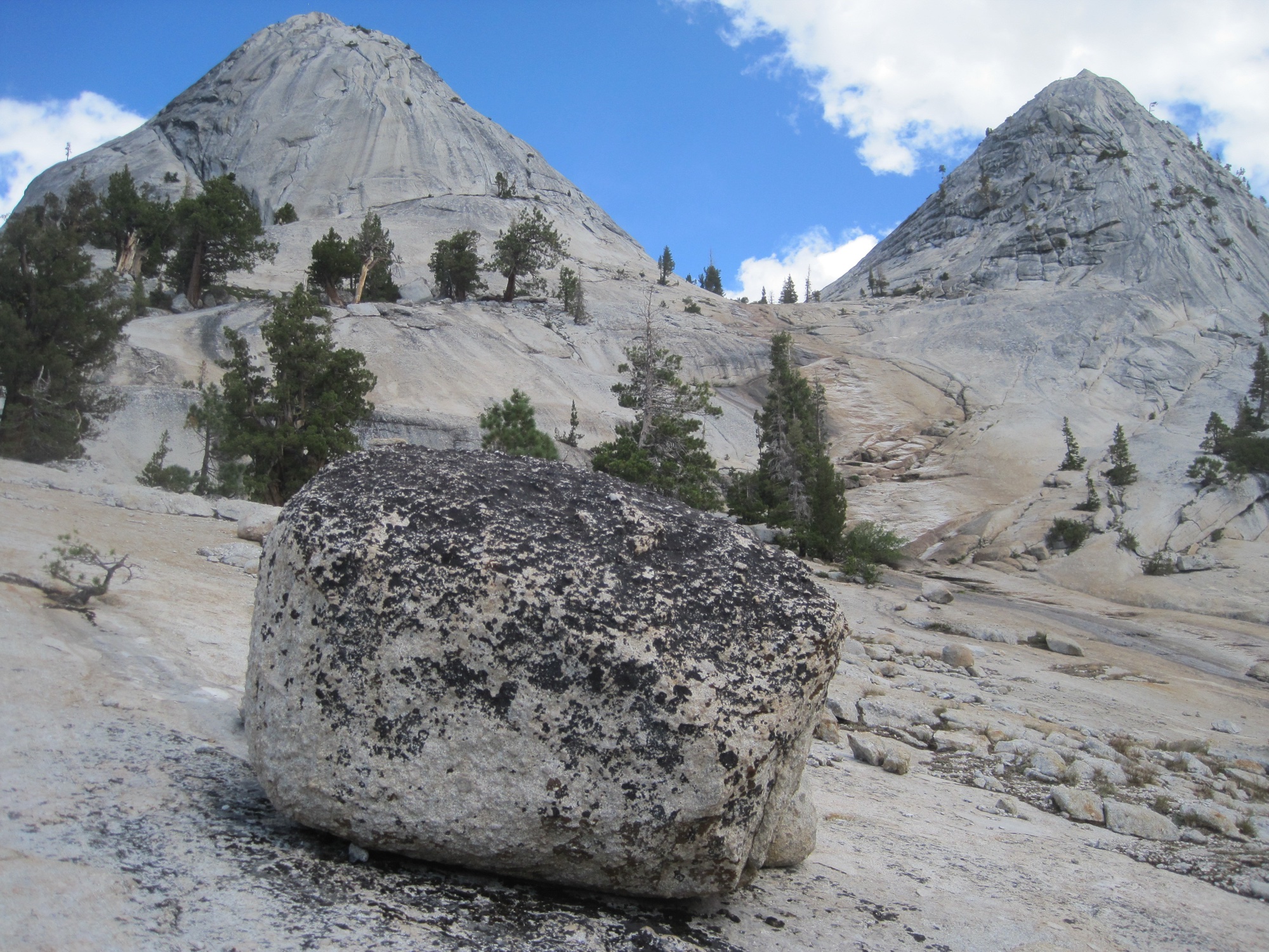 A lichen-spotted glacial erratic resting on bare granite below two domes